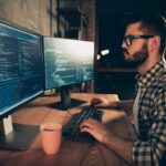 Close up side profile photo handsome he him his guy brainstorming briefing coder typing php css keyboard development outsource IT two monitors table office agency wear specs formalwear plaid shirt