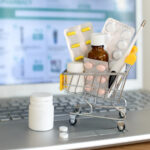 Shopping cart toy with medicaments in front of laptop screen with pharmacy web site on it. Pills, blister packs, medical bottles, thermometer set. Health care and internet shopping.